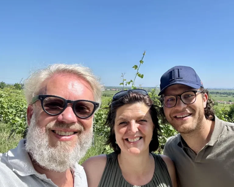Tre smilende personer tager en selfie i en grøn vinmark under en klar blå himmel.