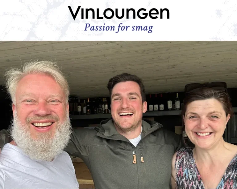 Tre smilende personer står tæt sammen i en vinbar med VinLoungen-logoet og sloganet “Passion for smag” øverst.
